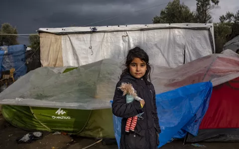 A girl holding her doll, standing in front of a tent in a migration encampment