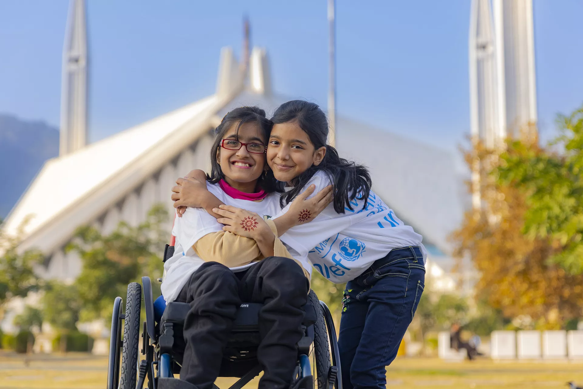Two girls hug in front of a mosque, one in a wheelchair